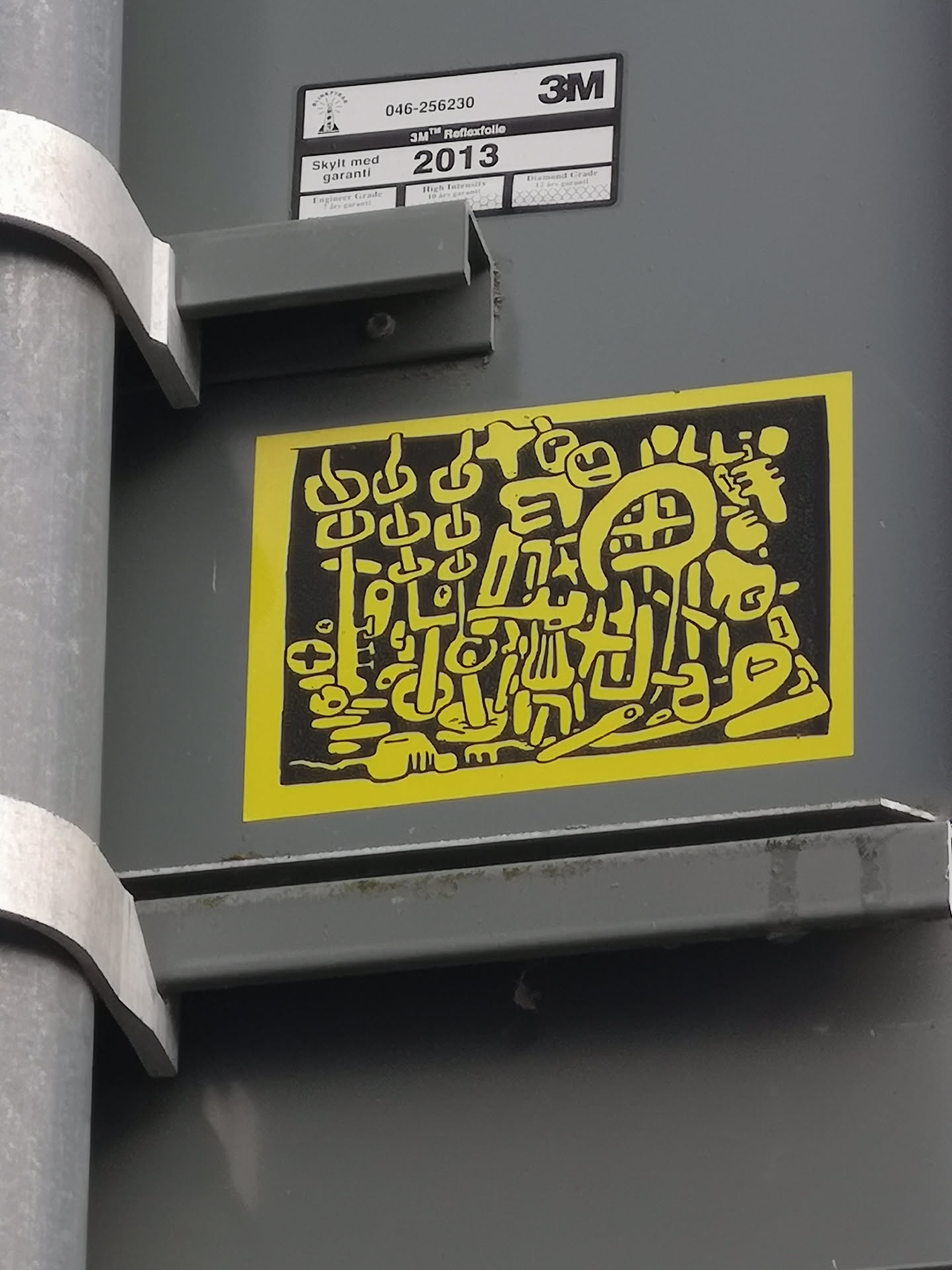 The sticker is a rectangular piece with a bright, almost neon yellow border. The interior is solid black, serving as a background for a dense, intricate pattern of yellow abstract shapes and lines. These shapes are organic and fluid, resembling a complex doodle or a collection of stylized symbols. Some elements suggest chains, others look like distorted faces, tools, or letters, all interwoven into a cohesive, yet unreadable, visual language. The overall impression is one of energetic, cryptic street art.