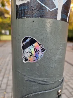 A holographic sticker featuring a stylized cartoon face with a bob haircut. The sticker is affixed to a weathered gray pole, which shows significant cracking and wear. The sticker's iridescent finish catches the light, making it stand out against the dull background.