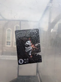 Street sticker A rectangular sticker on a wet glass surface, featuring a person in a black-and-white keffiyeh against a fiery background. It displays the text 'FREE GAZA' in a flame-styled font, a circular 'GLOBAL INTIFADA MOVEMENT' logo, and a QR code. The right side is partially torn.