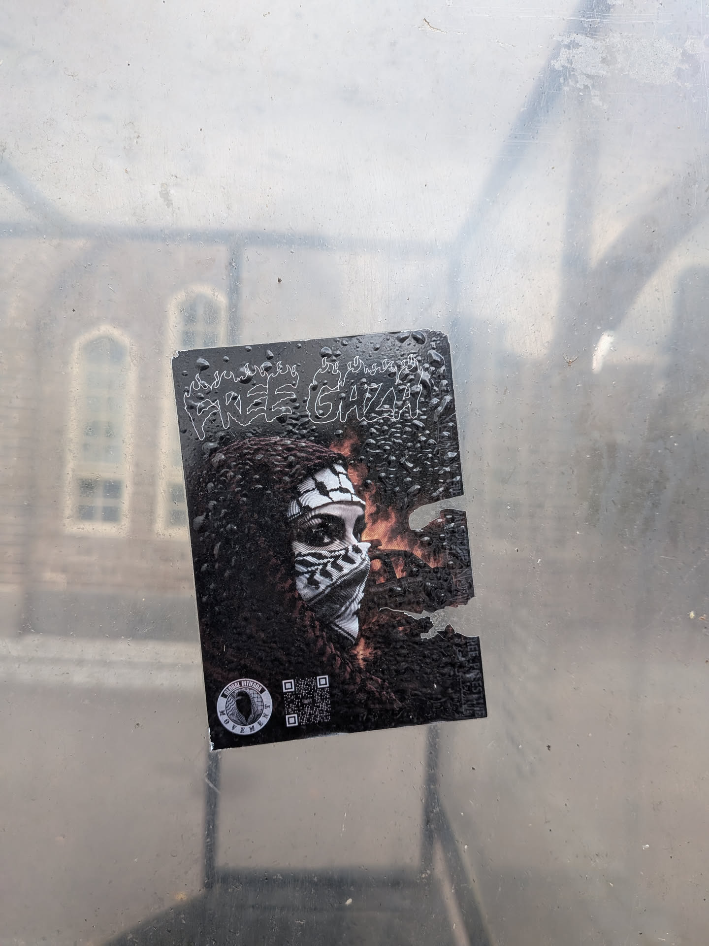 A rectangular sticker on a wet glass surface, featuring a person in a black-and-white keffiyeh against a fiery background. It displays the text 'FREE GAZA' in a flame-styled font, a circular 'GLOBAL INTIFADA MOVEMENT' logo, and a QR code. The right side is partially torn.