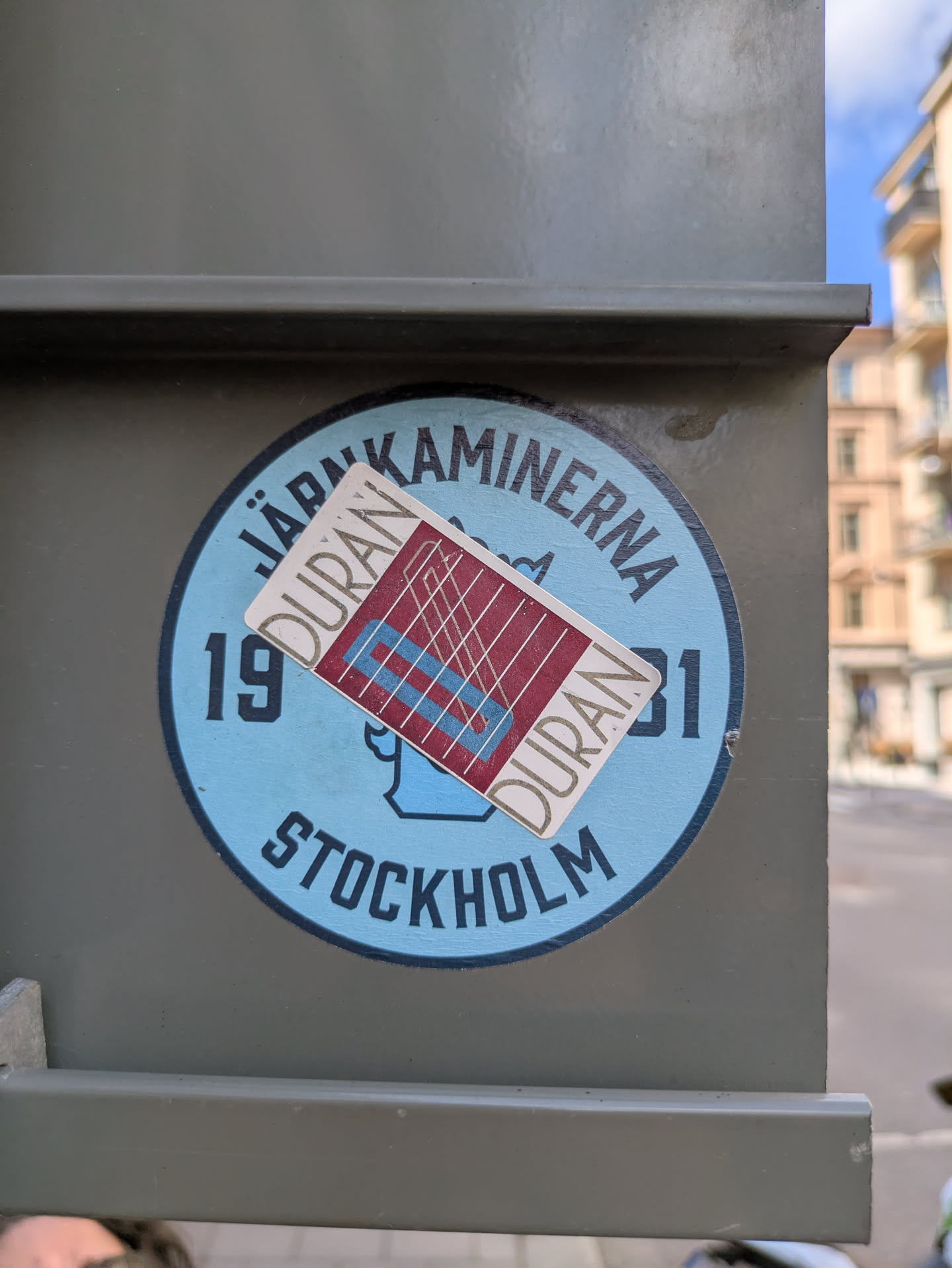 The central image features two distinct stickers overlaid on a dark olive-green utility box or pillar. The primary, larger sticker is circular with a light blue background and a dark blue outer border. Dark blue text arcs around the top, "JÄRNKAMINERNA," and bottom, "STOCKHOLM." The numbers "19" and "31" flank a stylized blue graphic of what appears to be a stove or chimney in the center. Partially covering this, at a slight angle, is a smaller, rectangular sticker with a white border. This smaller sticker has a maroon interior featuring a grid of thin white lines, two thick light blue diagonal lines, and two thick light blue horizontal lines. The word "DURAN" is written vertically in an outline font on both the left and right sides of this maroon section.
