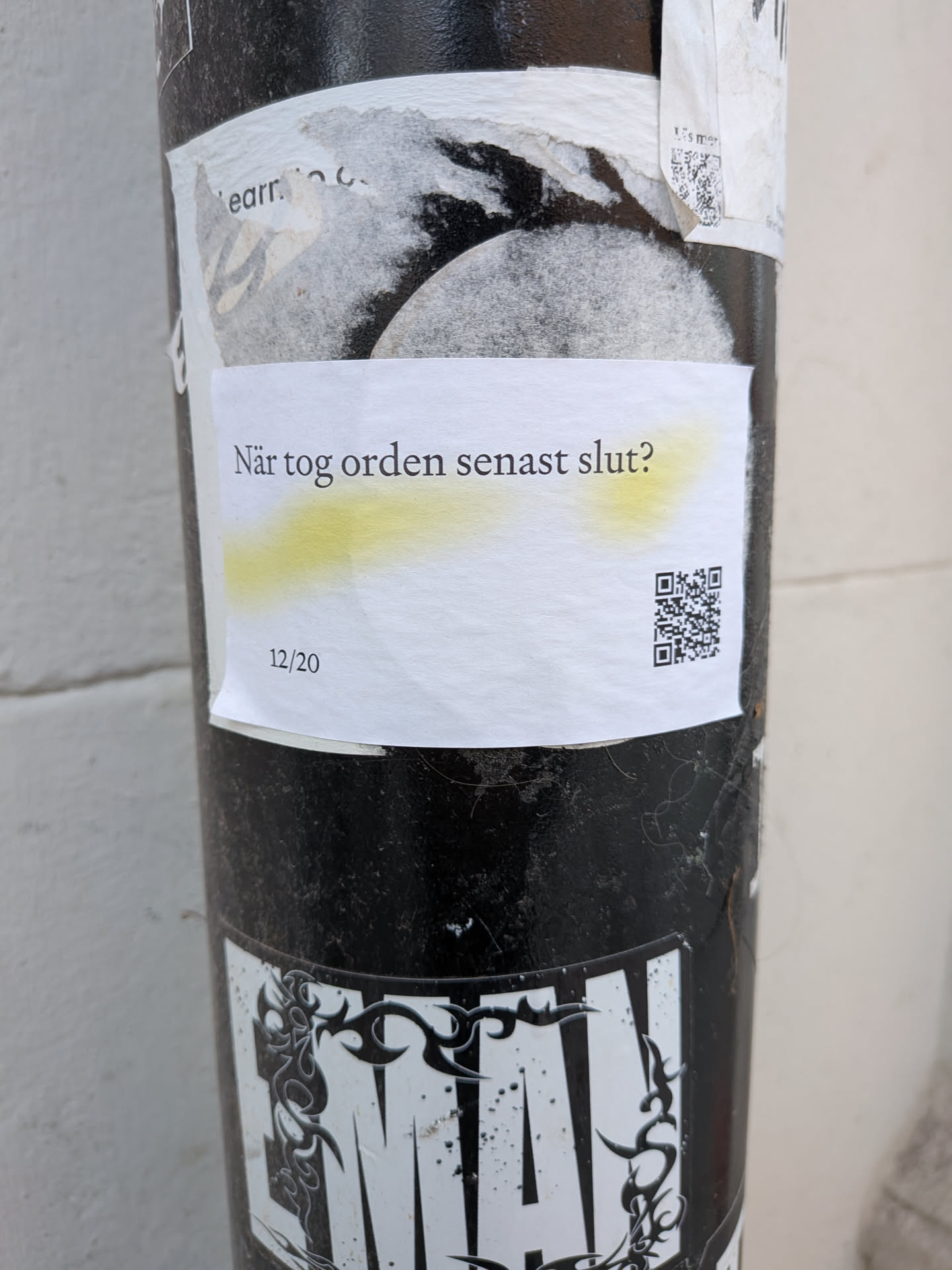 The sticker is a rectangular piece of white paper, slightly creased and textured, adhered to a dark, textured pole. It features black text in a sans-serif font. The main text, "När tog orden senast slut?", is centrally located and has a subtle, uneven yellow highlight underneath it. Below this, in the bottom left corner, are the numbers "12/20". In the bottom right corner, there is a clear, black and white QR code. The sticker appears to be placed over other older, torn stickers or papers, showing signs of wear and outdoor exposure.