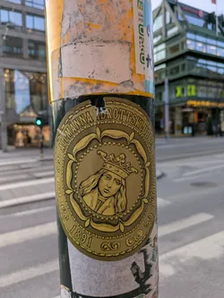 This round sticker features a gold design with an image of a crowned person, likely a historical figure,  and the text "Allmänna Idrottsklubben 1891". It appears to be affixed to a pole in an urban setting.