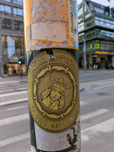 Street sticker This round sticker features a gold design with an image of a crowned person, likely a historical figure,  and the text "Allmänna Idrottsklubben 1891". It appears to be affixed to a pole in an urban setting.