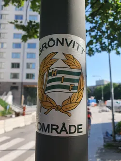 Street sticker A sticker featuring a logo with a green and white flag, gold wreath, and the text "GRÖNVITT" above and "OMRÅDE" below.  The letters HIF are visible on the flag.