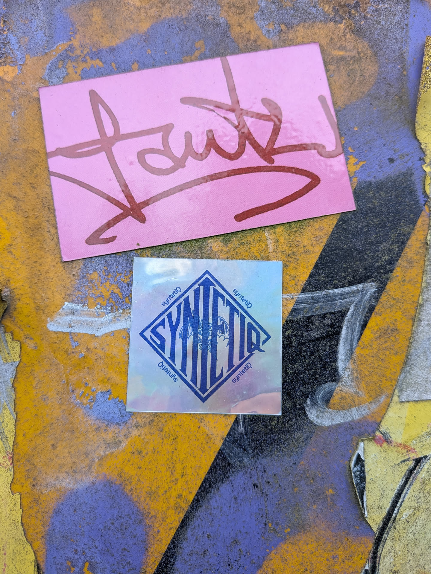 A square holographic sticker with a silver reflective background. It features a blue diamond containing the stylized text 'SYNTETIQ' and a faint winged graphic, surrounded by smaller 'syntetiq' text along its borders.
