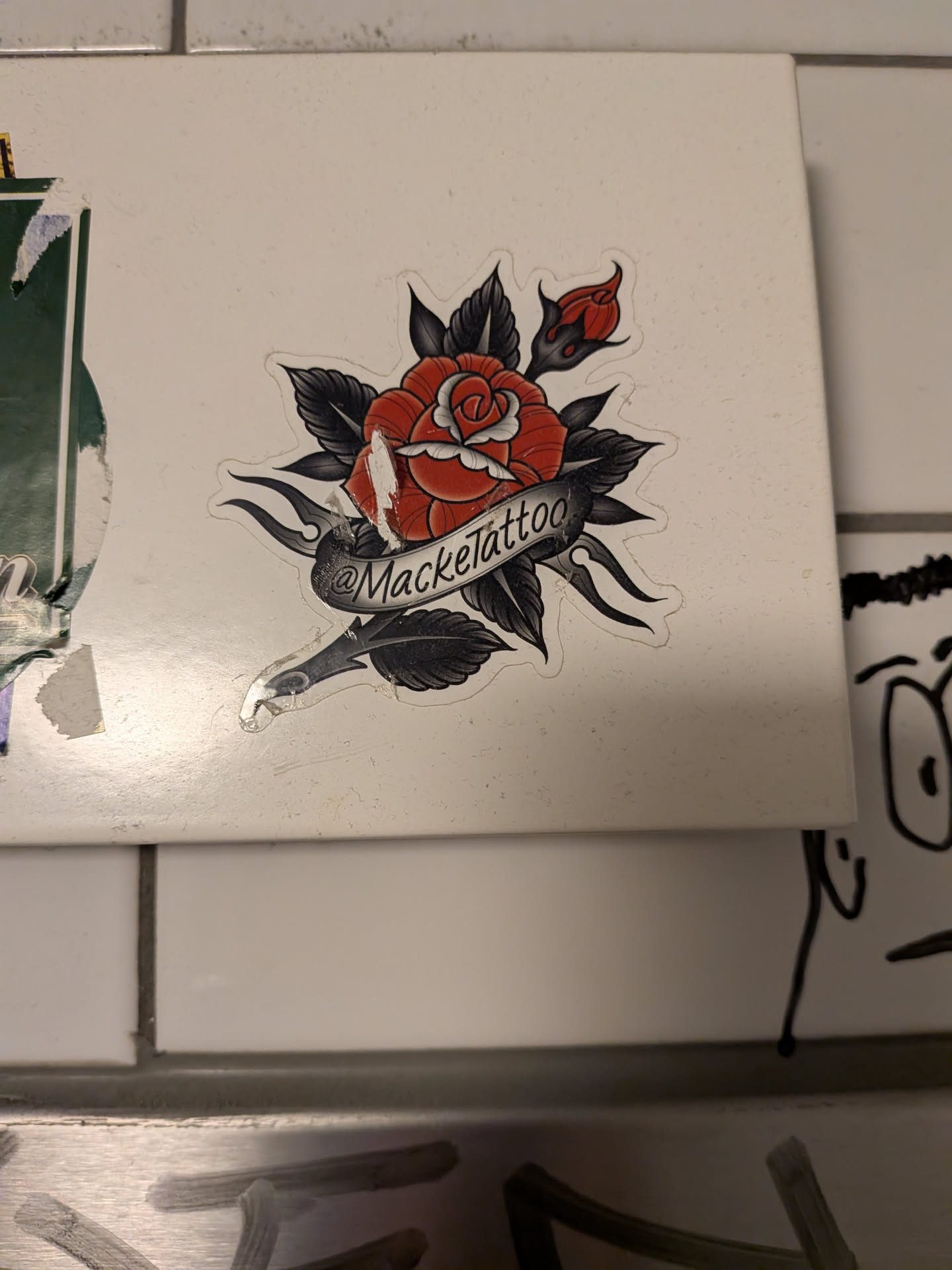 Street sticker The sticker features a traditional tattoo-style red rose with detailed petals and a bud. It is surrounded by dark, stylized leaves and flame-like accents. A banner wraps around the stem of the rose, bearing the text '@Macketattoo'. The sticker has a white border and appears to be slightly worn or damaged in places, particularly on the rose itself.