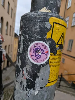 Street sticker The sticker is circular with a pink border and a light pink background. In the center, a cartoonish purple octopus is depicted. The octopus has large, wide eyes and is wearing a white mask with a bicycle symbol on it. It has eight tentacles, two of which are holding tools: a wrench and a screwdriver. One tentacle is also holding a small red object. The words "BIKE KITCHEN" are written in a circular fashion above the octopus, and "STUTTGART" is written below it, both in a purple, slightly stylized font.