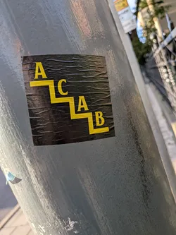 Street sticker A square black sticker with a wrinkled texture, adhered to a grey metal post. The sticker features the letters 'A', 'C', 'A', and 'B' in a yellow serif font, arranged sequentially down a yellow staircase-like line graphic from top-left to bottom-right.
