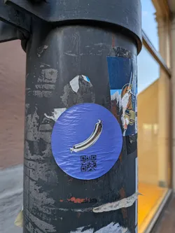Street sticker The sticker is circular with a vibrant blue background that has a subtle textured pattern, resembling ripples or waves. Centered on the sticker is a realistic illustration of a chrome-colored sausage, curved like a crescent moon. The sausage has a distinct metallic sheen and appears to be tied at one end. Below the sausage, a black QR code is printed, suggesting a link to further information or a website. The sticker is slightly creased, indicating it has been applied to a surface.