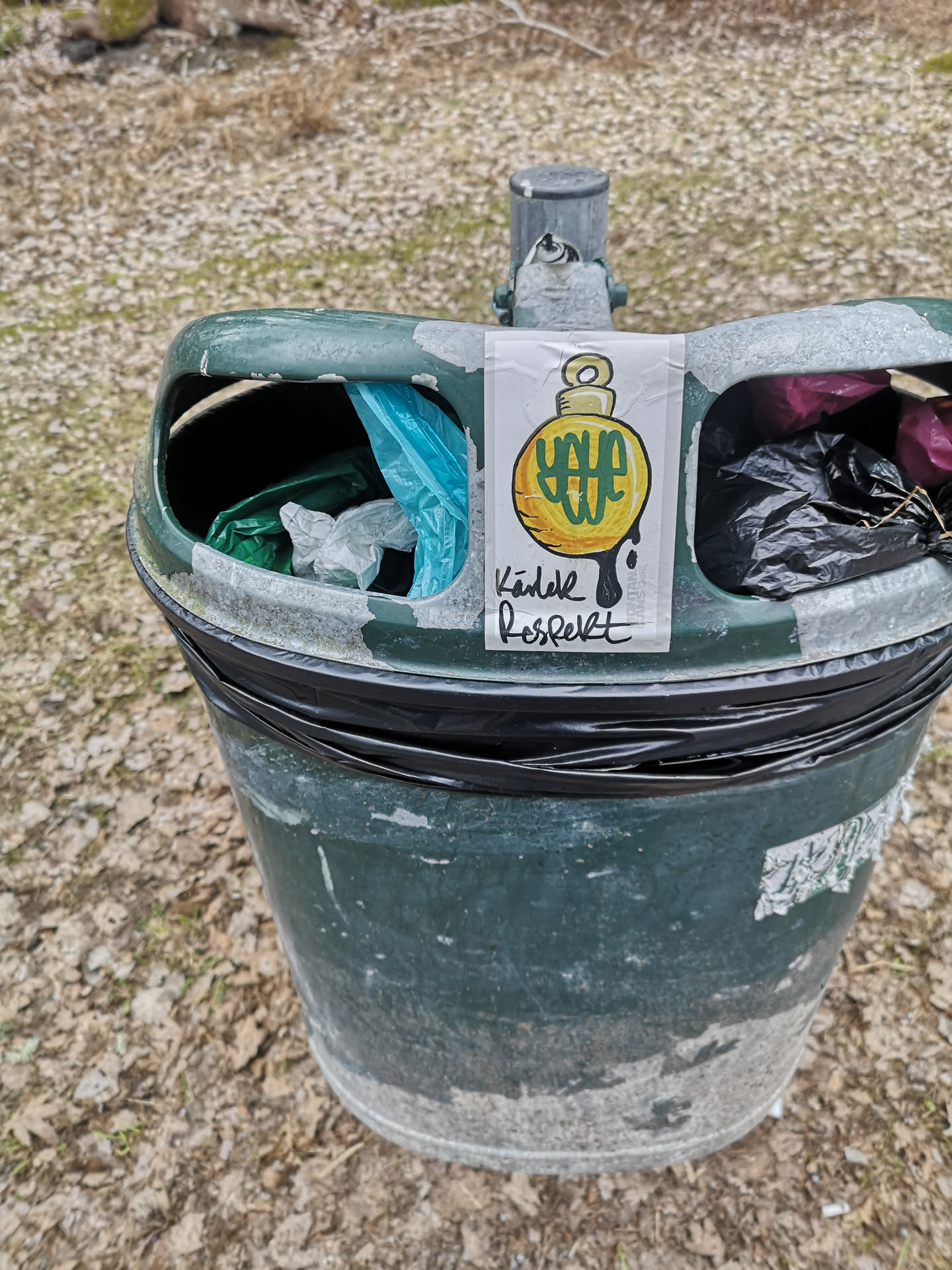 A rectangular white sticker featuring a central illustration of a stylized, round, yellow bomb-like object with a small, light yellow fuse/wick at the top. Inside the yellow bomb, there is a complex, stylized green symbol or text. Black drips extend downwards from the bottom of the yellow object. Below the illustration, handwritten-style text in black reads 'Kärlek Respekt'. The sticker is slightly wrinkled and adhered to a weathered, dark green trash can.