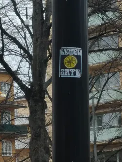 Street sticker The sticker is rectangular with a dark, likely black or very dark blue, background. In the center, there is a stylized, roughly drawn yellow lemon slice with visible segments. Above the lemon, the word "LEMON" is written in a light blue or faded white, blocky, somewhat distressed font. Below the lemon, the word "GATE" is written in a similar style and color. The entire design is framed by a thin, light blue or faded white border, giving it a somewhat grungy or street art aesthetic. The sticker appears to be slightly worn, with some edges and colors showing signs of age or exposure.