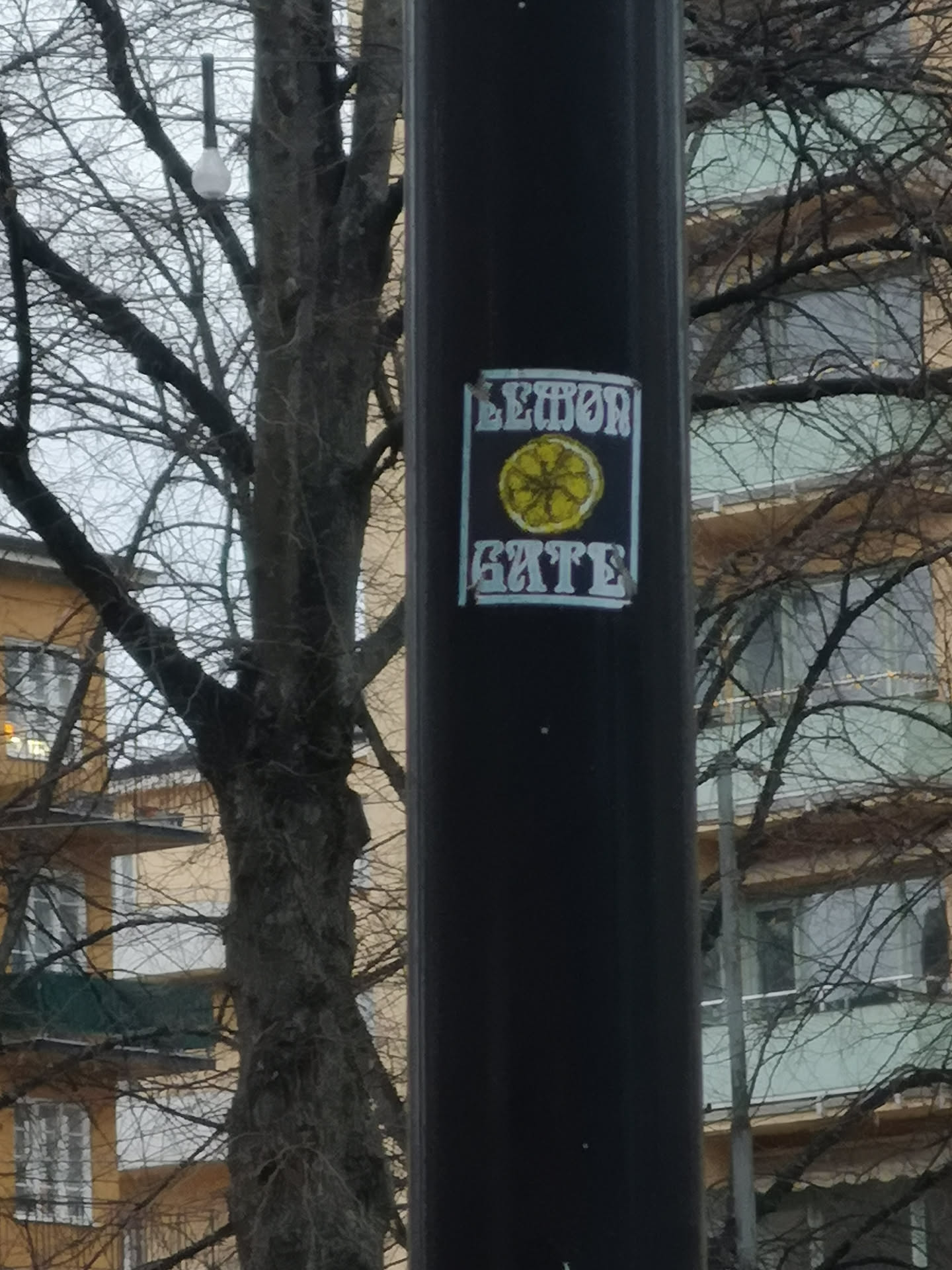 The sticker is rectangular with a dark, likely black or very dark blue, background. In the center, there is a stylized, roughly drawn yellow lemon slice with visible segments. Above the lemon, the word "LEMON" is written in a light blue or faded white, blocky, somewhat distressed font. Below the lemon, the word "GATE" is written in a similar style and color. The entire design is framed by a thin, light blue or faded white border, giving it a somewhat grungy or street art aesthetic. The sticker appears to be slightly worn, with some edges and colors showing signs of age or exposure.