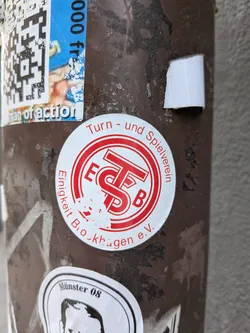 Street sticker The central sticker is circular, featuring a white background with red text and a red central emblem. The outer ring of the circle contains text: 'Turn - und Spielverein' curves along the top, and 'Einigkeit Brockhagen e.V.' curves along the bottom. The center of the sticker displays a stylized red logo composed of the letters 'E', 'T', 'S', and 'B', with 'E' and 'B' appearing to flank a larger 'TS' combination. The sticker shows signs of age and wear, with some of the red print slightly chipped or faded, particularly in the lower central area, and minor peeling around the edges. It is affixed to a dark brown, textured pole.
