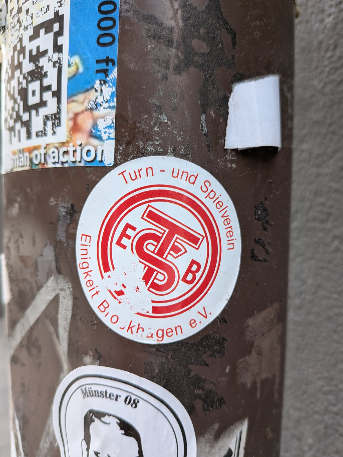 The central sticker is circular, featuring a white background with red text and a red central emblem. The outer ring of the circle contains text: 'Turn - und Spielverein' curves along the top, and 'Einigkeit Brockhagen e.V.' curves along the bottom. The center of the sticker displays a stylized red logo composed of the letters 'E', 'T', 'S', and 'B', with 'E' and 'B' appearing to flank a larger 'TS' combination. The sticker shows signs of age and wear, with some of the red print slightly chipped or faded, particularly in the lower central area, and minor peeling around the edges. It is affixed to a dark brown, textured pole.