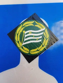 Street sticker A square sticker featuring a green and white flag with the letters HIF in the center, surrounded by a yellow laurel wreath. The sticker is adhered to a blue surface. The flag appears to be slightly damaged.