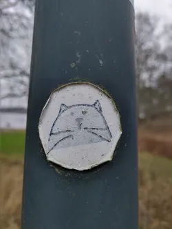 A small, round sticker featuring a simple illustration of a light grey cat. The style is minimalistic and charming. The sticker is affixed to a dark grey metal post outdoors.