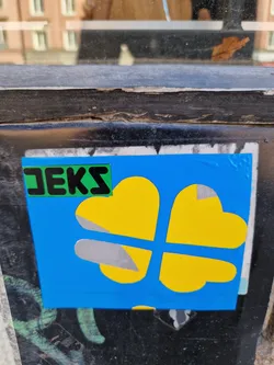 Close-up of a square sticker affixed to a dark surface. The sticker is predominantly blue, featuring a stylized yellow four-leaf clover.  A smaller green rectangle with the word "JEKS" written in black letters is visible on the sticker. 
