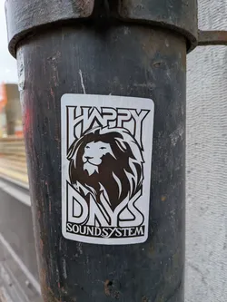 Street sticker The sticker is predominantly white with black graphics. It features a stylized, fierce-looking lion's head in the center, with a prominent mane that flows outwards. Above the lion's head, the word 'HAPPY' is written in large, bold, blocky capital letters with a distinctive outline. Below the lion's head, the letters 'DNY' are similarly styled, with the 'D' and 'Y' appearing larger and more prominent than the 'N'. At the very bottom, the words 'SOUND SYSTEM' are written in smaller, but still clear, capital letters. The overall design is sharp and graphic, with a strong contrast between the black and white elements.