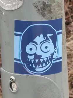 A square sticker featuring a stylized, cartoonish face in a monochromatic blue color scheme. The face is light blue with dark blue outlines and details. It has large, wide-open circular eyes with small pupils, and a wide, exaggerated grin revealing numerous sharp, triangular teeth. The hair is dark blue and appears somewhat messy. The background behind the face is light blue, framed by two horizontal dark blue stripes on either side. The sticker is applied to a grey, textured metal pole.