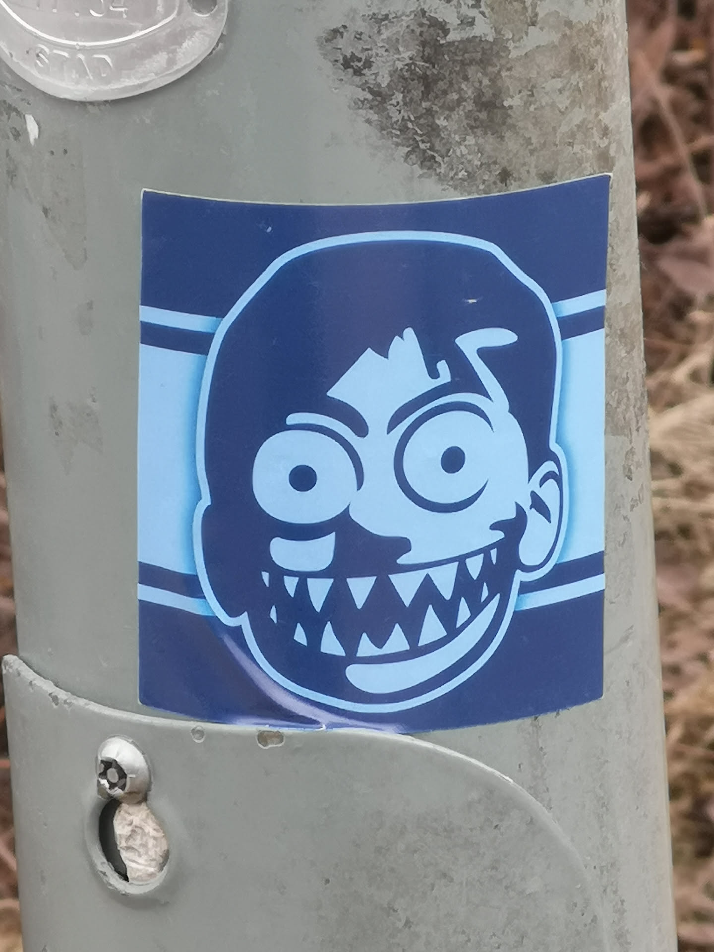 A square sticker featuring a stylized, cartoonish face in a monochromatic blue color scheme. The face is light blue with dark blue outlines and details. It has large, wide-open circular eyes with small pupils, and a wide, exaggerated grin revealing numerous sharp, triangular teeth. The hair is dark blue and appears somewhat messy. The background behind the face is light blue, framed by two horizontal dark blue stripes on either side. The sticker is applied to a grey, textured metal pole.