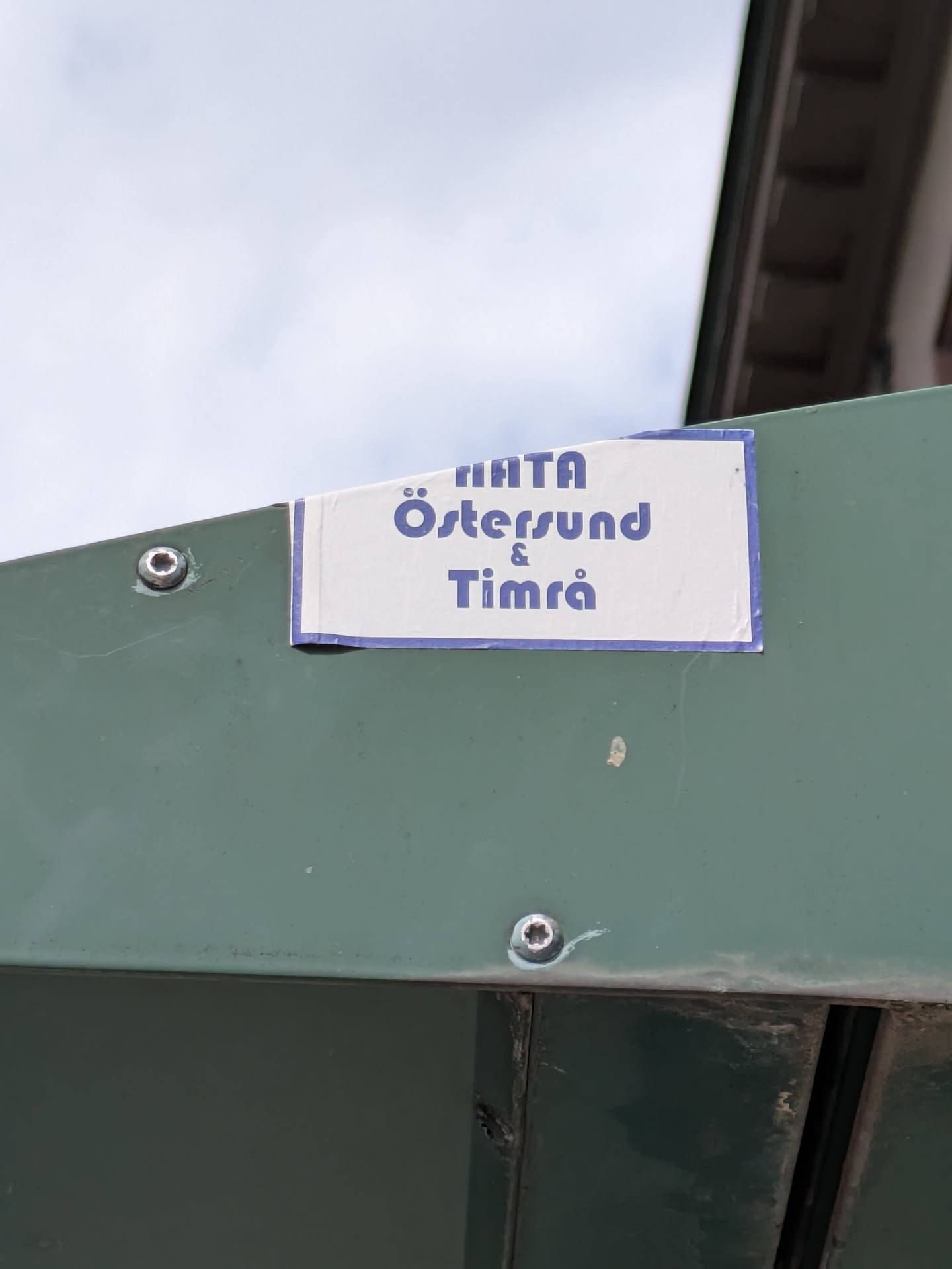 A rectangular white sticker with a blue border, attached to a green metal surface. The sticker displays the names "Östersund" and "Timrå" in blue text, possibly indicating support for these locations or the teams representing these locations.