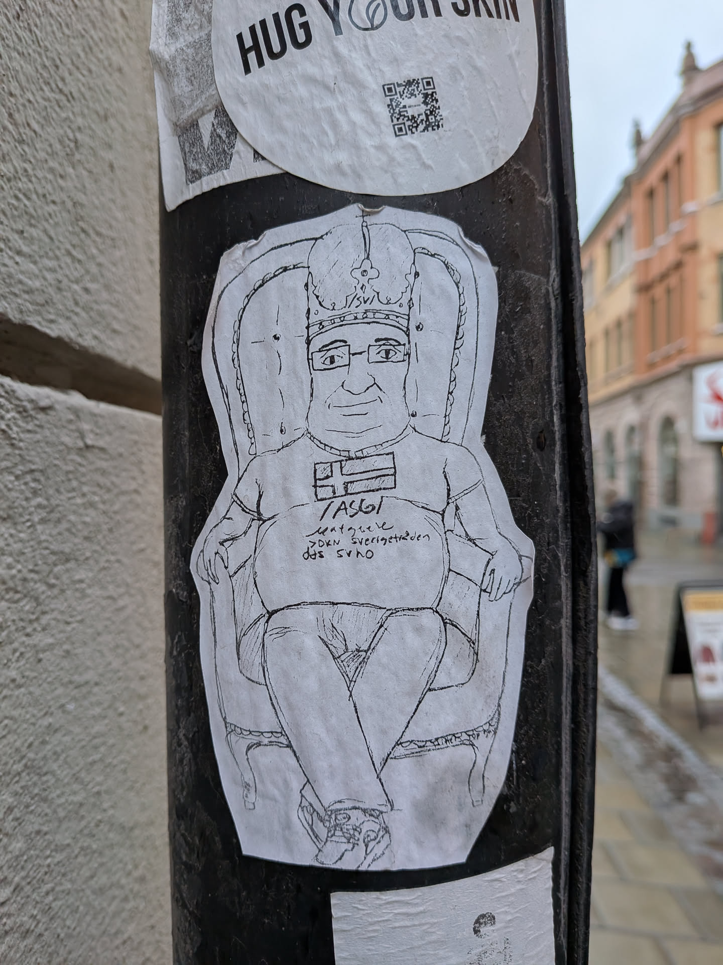 A monochrome, hand-drawn sticker featuring a portly, bespectacled man with a slight smile, wearing a crown (resembling a bishop's mitre) and a t-shirt adorned with the Swedish flag. He is depicted sitting in an ornate, high-backed armchair or throne, with his legs crossed. The drawing style is simple and cartoonish, rendered with black lines on white paper. The sticker has a slightly worn appearance with visible creases and edges.