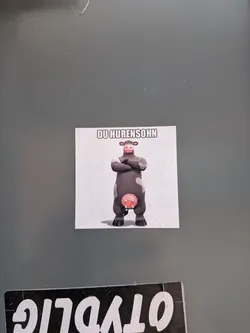 Street sticker A rectangular sticker featuring a cartoon cow, identifiable as Otis from the movie 'Barnyard', standing with its arms crossed. The cow is dark grey with white patches on its face and arms, and a prominent pink udder. It has a somewhat stern or confident expression. The background of the sticker is plain white, and the cow casts a subtle shadow beneath it. Above the cow, in bold, white block letters with a black outline, is the German phrase 'DU HURENSOHN'.