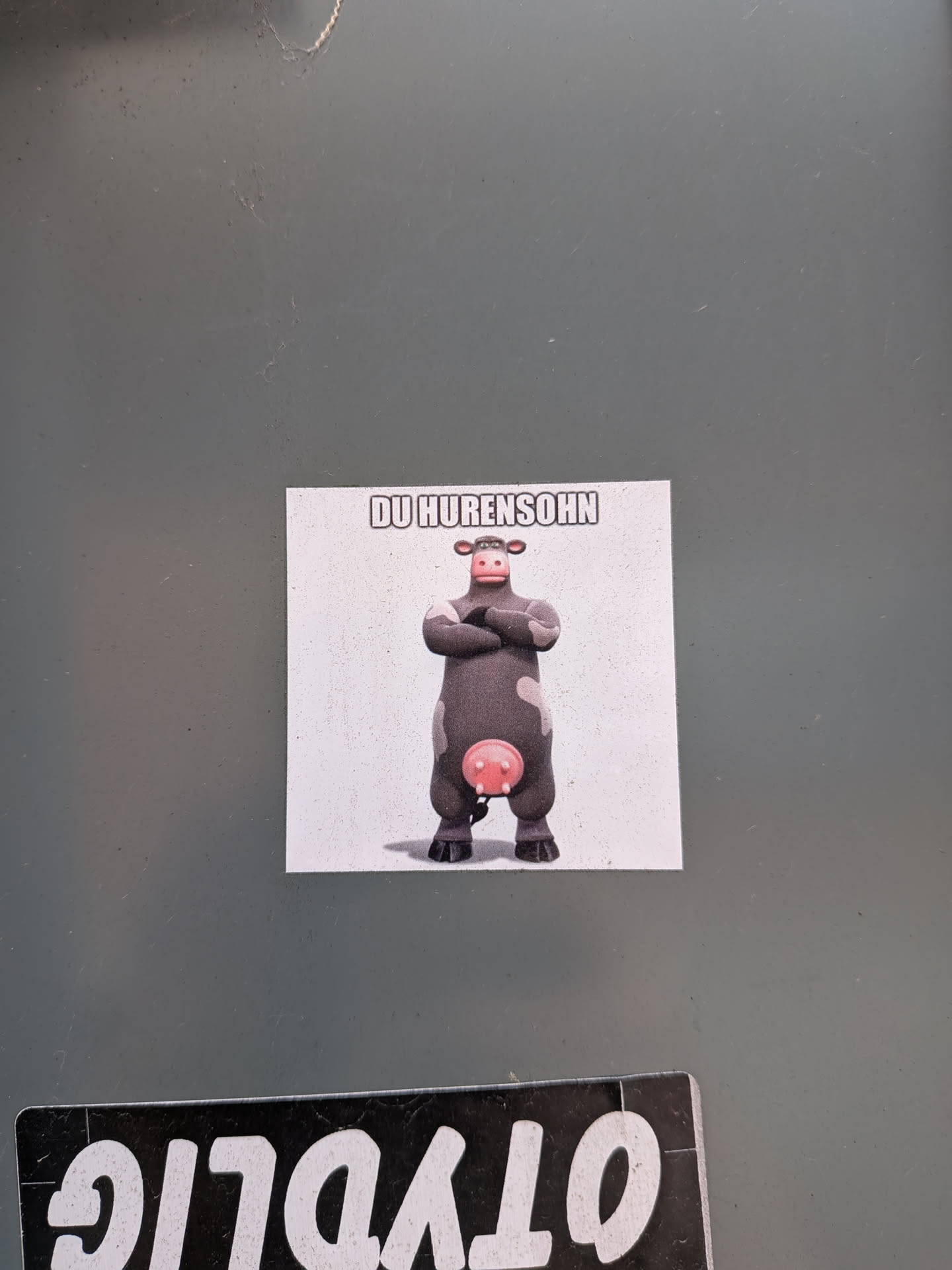 A rectangular sticker featuring a cartoon cow, identifiable as Otis from the movie 'Barnyard', standing with its arms crossed. The cow is dark grey with white patches on its face and arms, and a prominent pink udder. It has a somewhat stern or confident expression. The background of the sticker is plain white, and the cow casts a subtle shadow beneath it. Above the cow, in bold, white block letters with a black outline, is the German phrase 'DU HURENSOHN'.