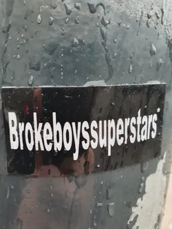 Street sticker A black sticker with the text "Brokeboysuperstars" in white, slightly reflective. It's affixed to a dark-colored, rain-wet surface.