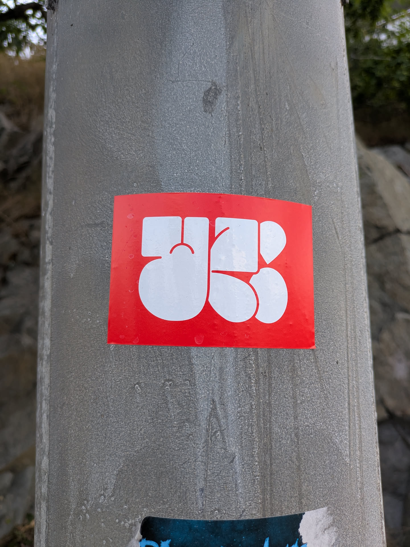 The sticker is a horizontally oriented rectangle with a vibrant red background. Centered on this background are large, interconnected white shapes that form a highly stylized, abstract symbol. The shapes are thick and rounded, creating a cohesive design that could be interpreted as a monogram or a unique emblem. The sticker is affixed to a textured grey metal pole and shows visible water droplets on its surface, indicating outdoor exposure.