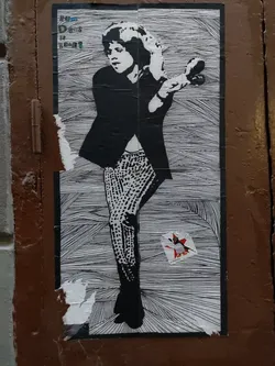 Street sticker A black and white street art sticker depicting Mick Jagger in a dynamic pose. The art style is reminiscent of a line drawing, with hatched lines creating shading and texture. The sticker is slightly damaged around the edges, suggesting it has been affixed to a surface for some time.  A smaller sticker is visible underneath the main one.