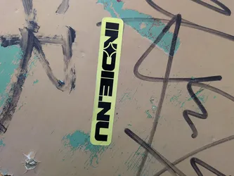 Street sticker A vertical, rectangular sticker with rounded corners, featuring a vibrant lime green background. The text 'INDIE.NU' is prominently displayed in bold black capital letters. The letter 'E' is uniquely stylized, incorporating a star or diamond shape within its negative space, giving it a distinctive graphic element. The sticker is applied to a textured, beige-green surface with black graffiti marks.