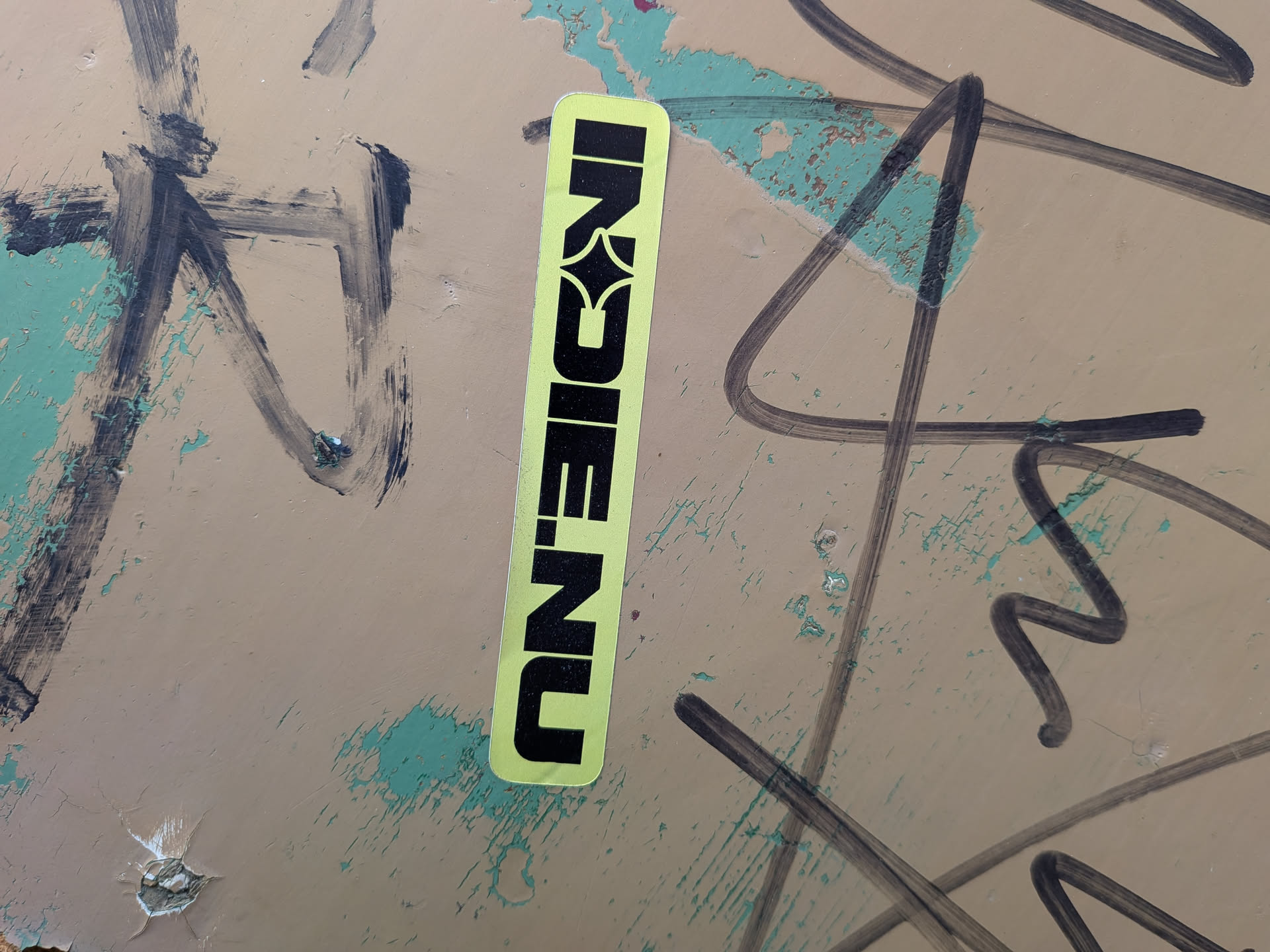 A vertical, rectangular sticker with rounded corners, featuring a vibrant lime green background. The text 'INDIE.NU' is prominently displayed in bold black capital letters. The letter 'E' is uniquely stylized, incorporating a star or diamond shape within its negative space, giving it a distinctive graphic element. The sticker is applied to a textured, beige-green surface with black graffiti marks.