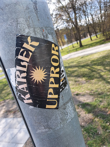 Street sticker Close-up of a worn sticker affixed to a metal pole. The sticker is primarily black with yellow/gold text that reads "KARISIER UPPPROR." A stylized sun is centered on the sticker. The background shows a blurred park-like setting with trees and grass.