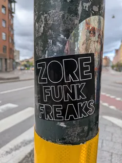 Street sticker A rectangular sticker with a solid black background featuring three lines of text in bold, white outlined, sans-serif capital letters. The text reads 'ZORE' on the first line, 'FUNK' on the second, and 'FREAKS' on the third. The sticker is applied to a dark, textured utility pole, slightly angled, with its edges clearly visible.