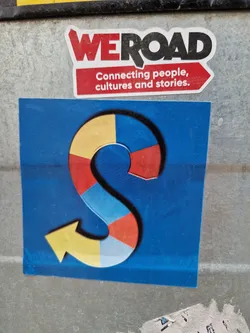 Street sticker A sticker featuring a colorful arrow shaped as an S on a blue background. The upper sticker says: WE ROAD Connecting people, cultures and stories. The design suggests a journey, connection between cultures, and sharing of stories.