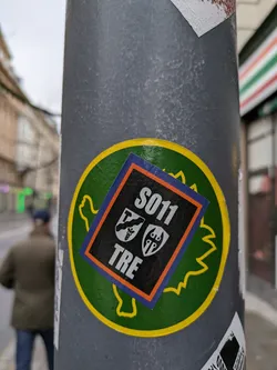 Street sticker A black square sticker with the text S011 and TRE, along with two crest-like emblems, affixed to a larger round green sticker shaped like a stylized leaf or creature. The larger sticker is attached to a grey pole.