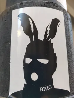 Street sticker The sticker features a minimalist design of a person wearing a balaclava and bunny ears, creating a stark contrast between the anonymous mask and the playful ears. The artist's signature or brand name is "BRIO", subtly placed at the bottom.