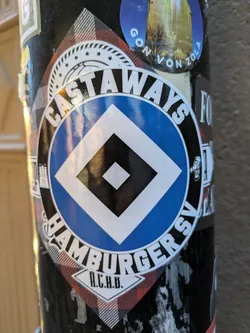 Street sticker The sticker features a circular logo for Hamburger SV, a German sports club. The prominent element is a blue circle with a white diamond shape inside, bordered by a thin black line. Surrounding the blue circle is a white ring with black text that reads "CASTAWAYS HAMBURGER SV". Below this, on a white diamond shape with red and black plaid accents, are the letters "A.C.H.B.". The sticker appears to be circular overall, with some white decorative elements like a border of small stars above the main logo.