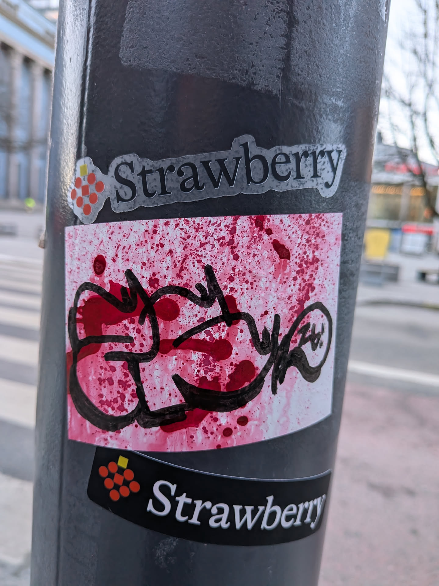 The central sticker is a rectangular piece with a white background heavily splattered with various shades of red, ranging from bright crimson to darker, almost maroon spots. The splatters are unevenly distributed, creating a dynamic and somewhat chaotic texture. Overlaid on this red-splattered canvas is a prominent, thick black graffiti tag or script. The lines of the tag are fluid, interconnected, and highly stylized, forming an illegible but energetic word or symbol. The black ink appears to be applied directly over the red splatters, giving it a layered effect. The overall appearance is raw, energetic, and embodies a street art aesthetic.