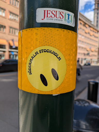 A round yellow sticker on a green pole features a smiley face with simple oval eyes and no mouth. The words "Södermalm Stockholm" are written in black around the top half of the circle. The sticker has a reflective, honeycomb-like background. Above the sticker is another sticker saying "Jesus loves people" with an image containing a cross and menorah.