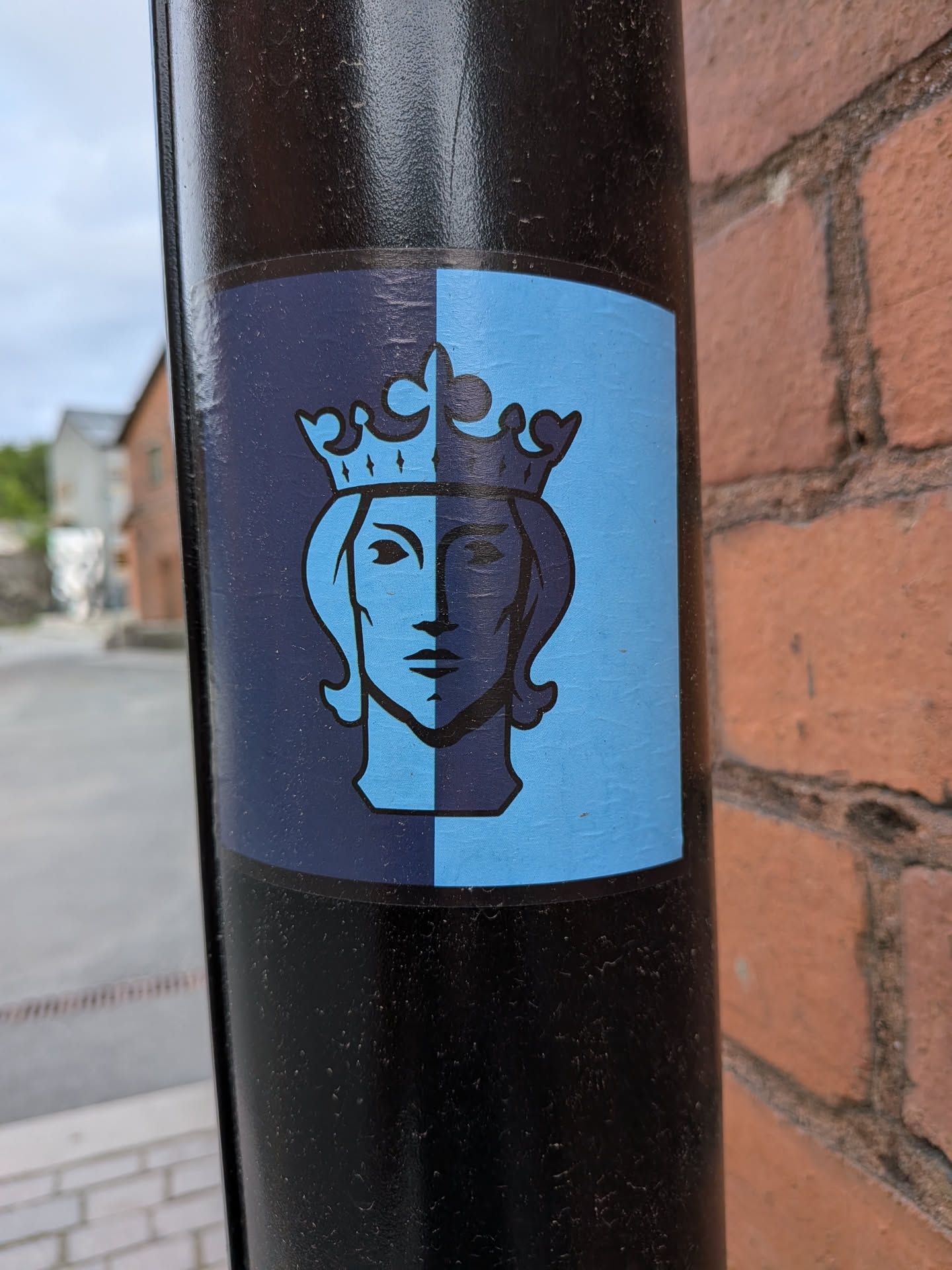 The sticker features a stylized depiction of a human head wearing a crown, presented in a rectangular format. The background of the sticker is vertically split: the left half is dark navy blue, and the right half is a lighter sky blue. The central image of the crowned head is also split in color, mirroring the background: the left side of the head (on the dark navy background) is light sky blue, and the right side of the head (on the light sky blue background) is dark navy blue. All outlines of the head and crown are rendered in black. The design has a clean, iconic, and somewhat symmetrical appearance, reminiscent of a heraldic crest or emblem. The sticker is applied to a textured black pole.