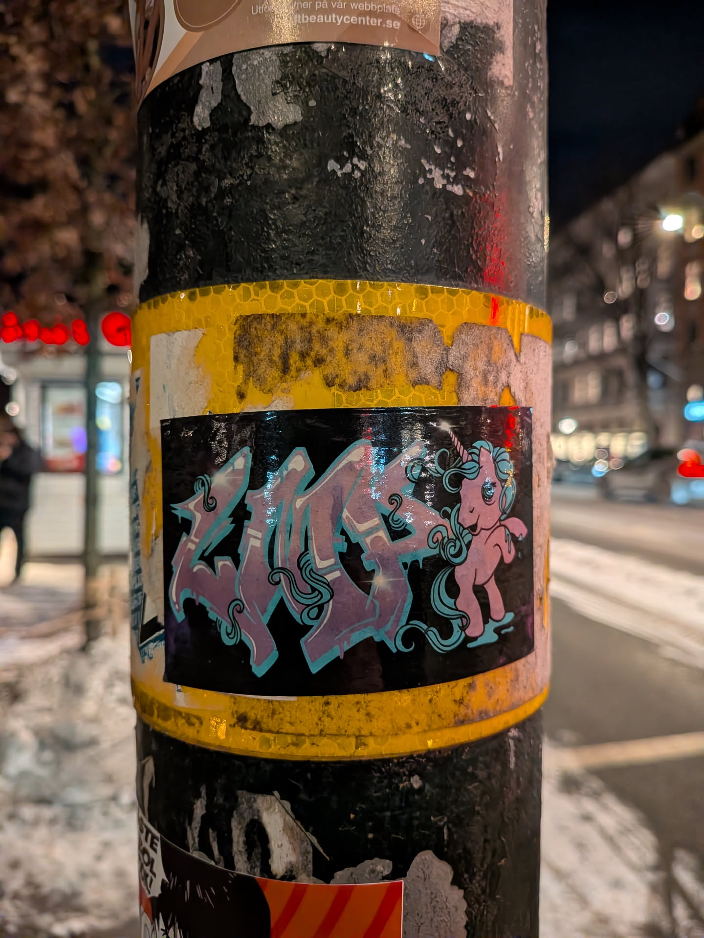 Street sticker The sticker features a glossy black background. On the left, stylized graffiti text, likely 'ZAP!', is rendered in shades of purple with light blue outlines and highlights, giving it a bubbly, dynamic appearance. To the right, a cute, cartoon-style pink unicorn stands, facing left towards the text. The unicorn has a light blue mane and tail, a small horn, and a playful expression. The overall style is bright and energetic, contrasting the dark background.