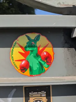 This round sticker features a green kangaroo wearing red boxing gloves, set against a background of yellow and red rays. The text "JOHNBEJER.COM" is curved around the left side of the design.