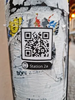Street sticker A white square sticker with a QR code and the text "Station 2a" below it. The sticker is affixed to a pole that is covered in other stickers and graffiti.
