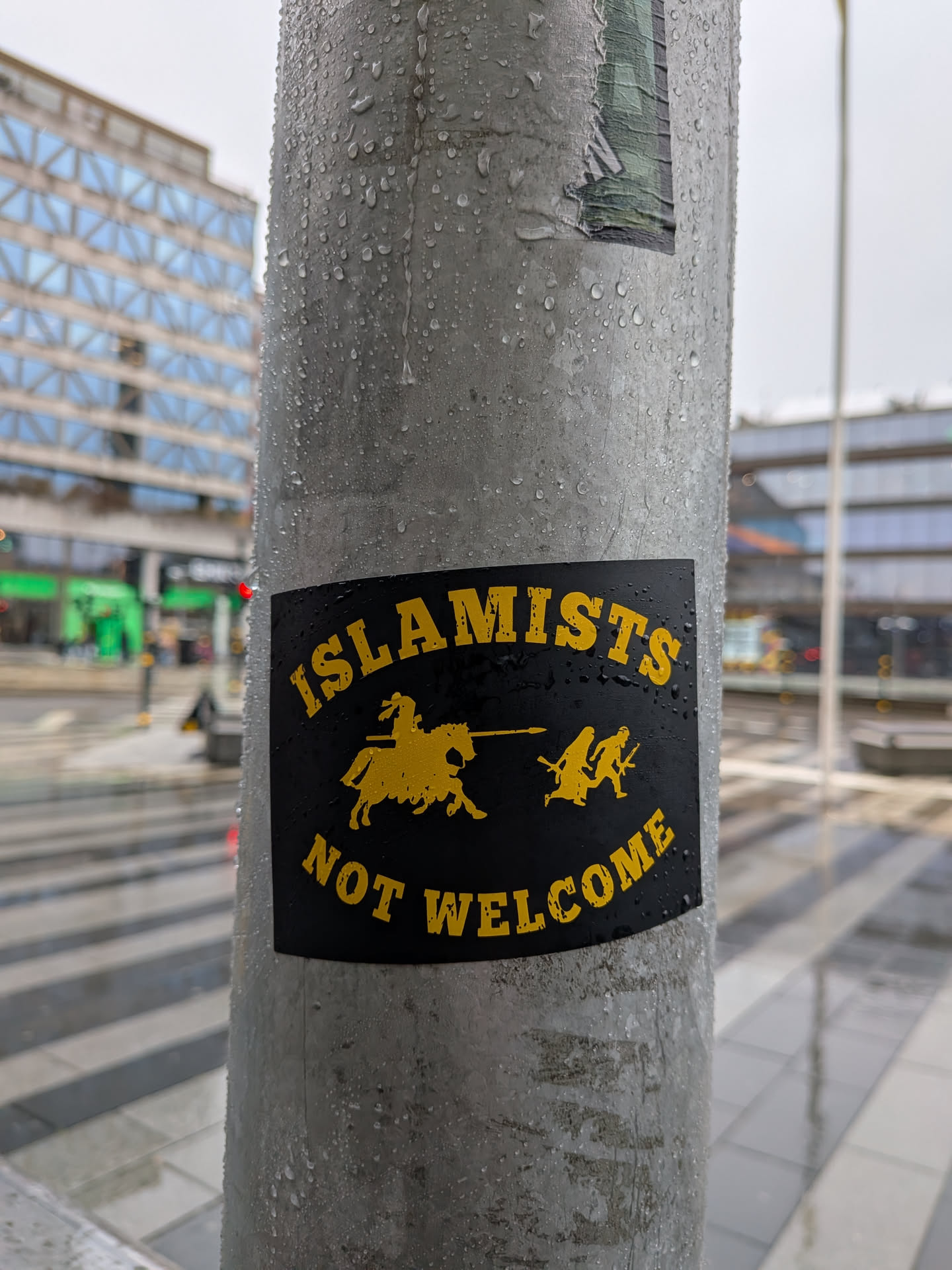 The sticker is rectangular with a black background and all elements rendered in bright yellow. The top text reads 'ISLAMISTS' in an arched font. Below this, a central image depicts a silhouette of a knight on horseback, holding a lance, charging towards two smaller, silhouetted figures who appear to be running or fleeing. The bottom text, also arched, reads 'NOT WELCOME'. The overall design is stark and graphic, reminiscent of historical or political propaganda.
