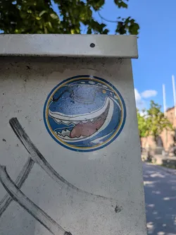 Street sticker A round sticker featuring a stylized blue shark-like monster with an open mouth, sharp teeth, and a pink tongue, framed by a blue and yellow border.