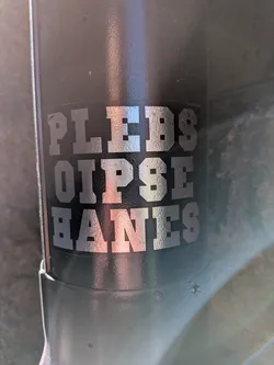 Street sticker The sticker features three lines of text: 'PLEBS' on the top, 'OIPSE' in the middle, and 'BANES' at the bottom. The letters are rendered in a bold, blocky, sans-serif font with a slightly distressed or textured appearance, giving them an industrial or collegiate feel. The text is a metallic silver-grey color, reflecting light, and is set against a dark, possibly black or very dark grey, background. The sticker appears to be a vinyl decal applied to a textured, dark surface, possibly a water bottle or similar item, with some minor wear visible on the edges of the letters.