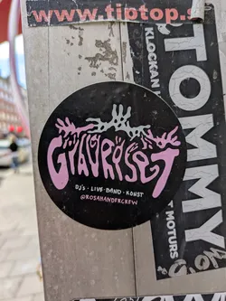 Street sticker This is a round sticker with a black background. The main text 'GYAVRYSET' is prominently displayed in a decorative, stylized font in pink. Above the text, there's a white abstract graphic. Below the main text, it says 'DJ'S . LIVE-BAND . KONS' and the Instagram handle '@ROSAHANDERCREW'.